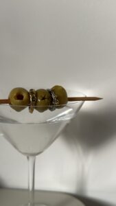 A martini glass filled with green olives, with cocktail picks resting across the top and several rings visible on the olives.