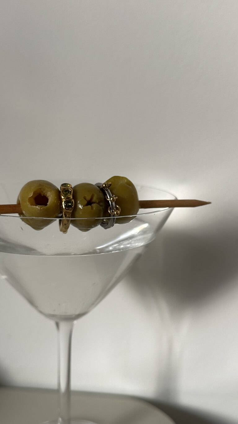A martini glass filled with green olives, with cocktail picks resting across the top and several rings visible on the olives.