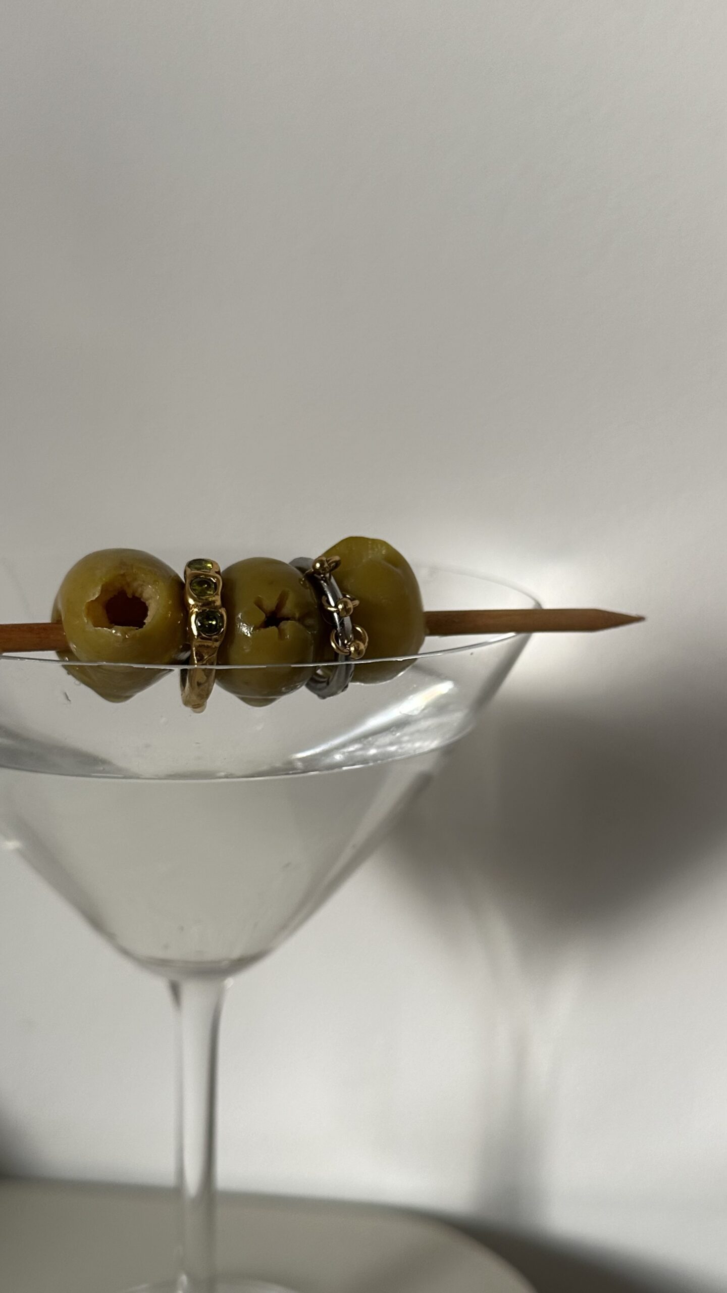 A martini glass filled with green olives, with cocktail picks resting across the top and several rings visible on the olives.