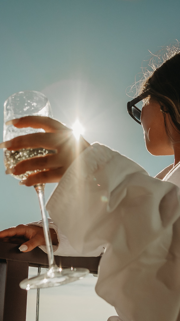 A woman in sunglasses holds a glass of wine outdoors, with sunlight shining through the glass against a clear blue sky.