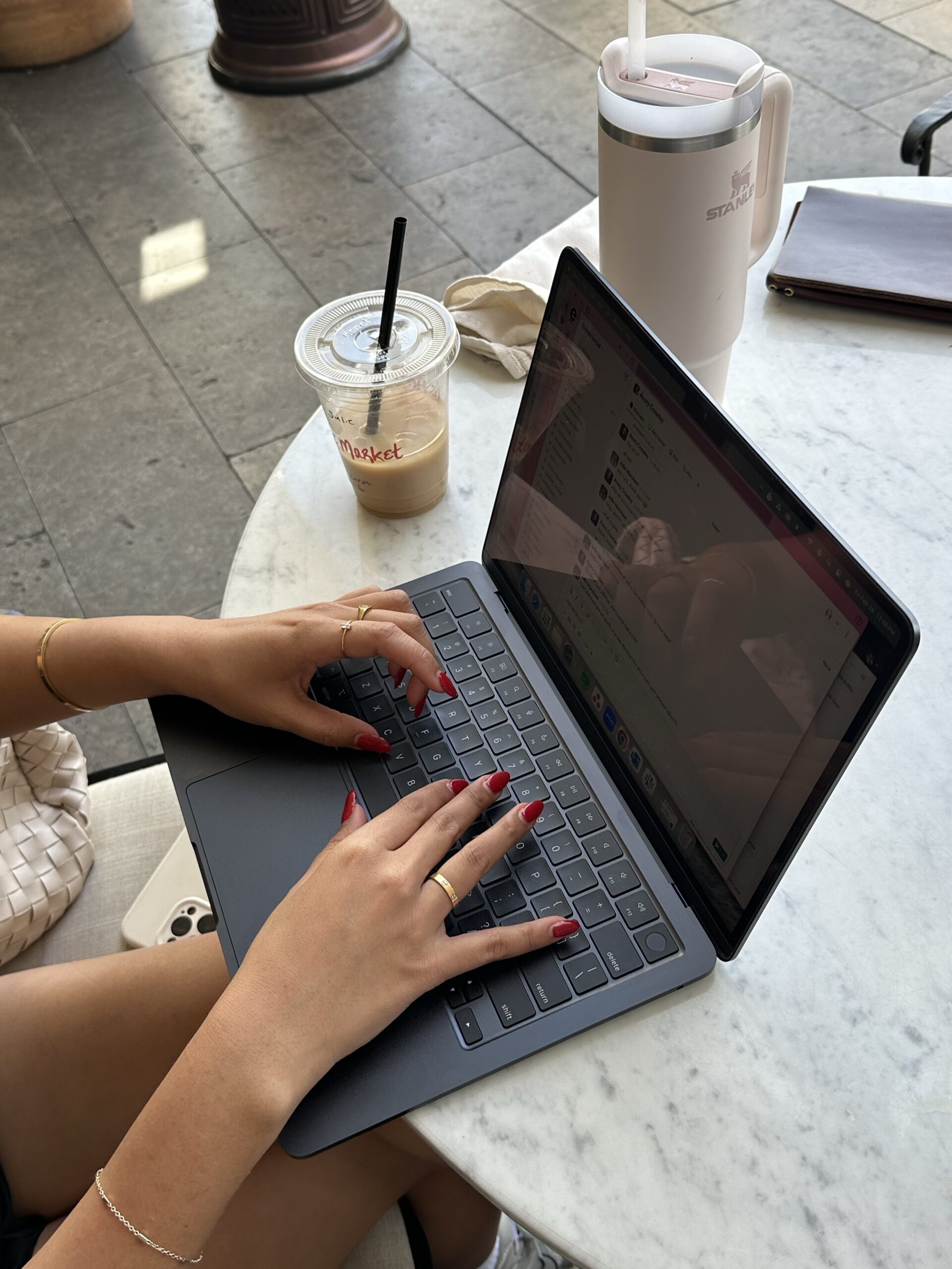 A person types on a laptop at a marble café table, with an iced coffee, reusable tumbler, and notebook nearby.