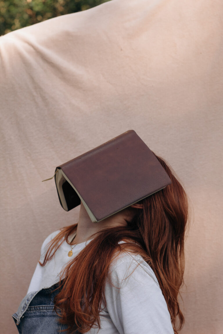 Woman with long red hair wearing a white shirt and denim overalls, covering her face with a book while leaning against a neutral beige wall.